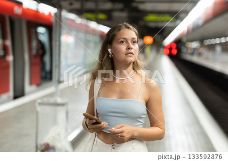 Girl with earphones and phone is standing on subway platform 133592876