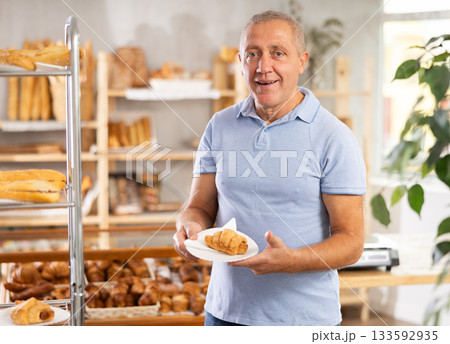 Senior male customer stands with croissant in hands near window of bakery. 133592935
