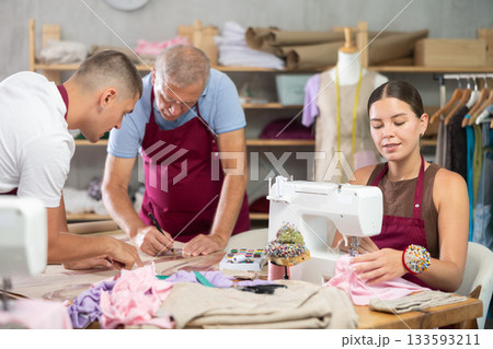 Girl master sew clothes in sewing machine, senior craftsman with guy helper draw dress pattern Girl master sew clothes in sewing machine, senior craftsman with guy helper draw dress pattern 133593211