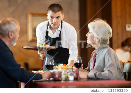 Waiter brings order to elderly man and woman couple Waiter brings order to elderly man and woman couple 133593390