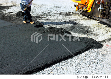 Construction worker spreads fresh asphalt on ground while paving machinery operates Construction worker spreads fresh asphalt on ground while paving machinery operates 133594811