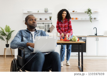 A man in a wheelchair works on a laptop while his partner prepares food in a modern kitchen. 133597839