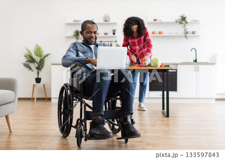 A man in a wheelchair uses a laptop while his partner prepares food in the kitchen. 133597843