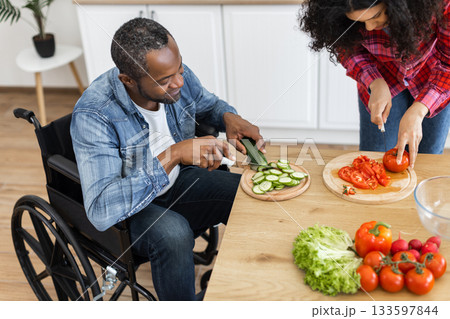 A diverse couple prepares a healthy meal together in a bright kitchen, one using a wheelchair. A diverse couple prepares a healthy meal together in a bright kitchen, one using a wheelchair. 133597844