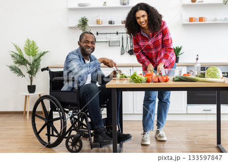 A smiling couple prepares a meal together in a bright, modern kitchen, with one person using a wheelchair. A smiling couple prepares a meal together in a bright, modern kitchen, with one person using a wheelchair. 133597847