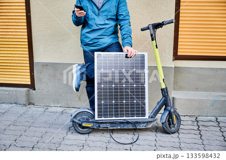 Cropped view of male person holds solar panel, for charging electric scooter, using smartphone. Integration of solar power as sustainable energy source, promoting eco-friendly urban transportation. Cropped view of male person holds solar panel, for charging electric scooter, using smartphone. Integration of solar power as sustainable energy source, promoting eco-friendly urban transportation. 133598432