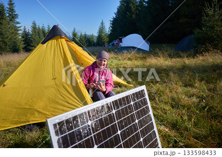 Happy woman sits in front of yellow tent, charging her phone by using solar panel, highlighting use of renewable energy and eco-friendly camping. On background lush green trees and clear blue sky. 133598433
