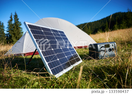 Tourist tent with solar panel and portable power station nearby in grassy field. Scene surrounded by rolling hills, tall pine trees under clear blue sky, showcasing eco-friendly camping setup. 133598437