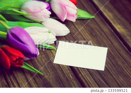 Flat design is showing multicolored tulip cluster on wooden slatted table with blank white card Flat design is showing multicolored tulip cluster on wooden slatted table with blank white card 133599521