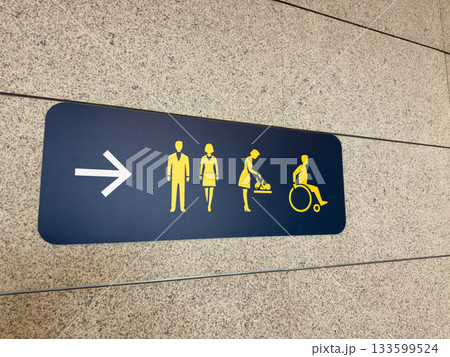 Directional signs on wall for restrooms for men and women, baby changing room, and wheelchair accessible toilet. Wall sign with arrow indicates direction of public WC 133599524