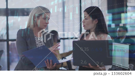 Discussing market data businesswomen holding binder and laptop at trading desk, with ticker overlay 133602604