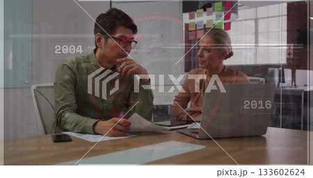 Two colleagues wearing business attire reviewing documents and laptop at office table, with notes 133602624