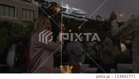 Waving sparklers around Greek flagpole friends in bomber jacket on patio, with string lights 133602771