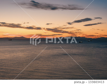 Sunset over calm waters with fishing ships on the horizon near coastal mountains at dusk 133603413