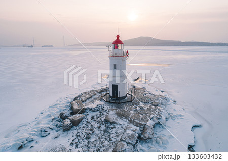 Lighthouse standing on frozen shore in Vladivostok during winter sunrise with ships in the distance Lighthouse standing on frozen shore in Vladivostok during winter sunrise with ships in the distance 133603432