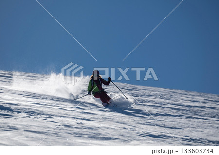 A female skier carves through fresh powder on a sunny mountain slope, kicking up snow as she descends, creating a dynamic and energetic winter sports action scene under a clear blue sky 133603734