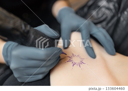 close-up of a tattoo artist outlining a sun design on a client skin, showing the precision of the tattoo process, sterile gloves, and professional equipment. close-up of a tattoo artist outlining a sun design on a client skin, showing the precision of the tattoo process, sterile gloves, and professional equipment. 133604460