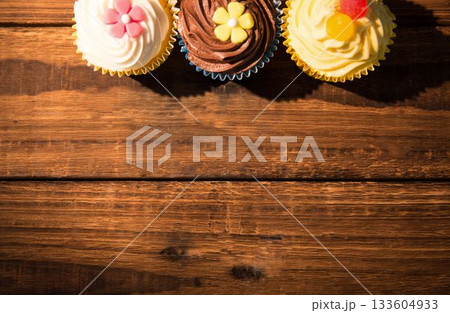 Three frosted cupcakes are resting on wood grain table in flat design with fondant flowers, gummies Three frosted cupcakes are resting on wood grain table in flat design with fondant flowers, gummies 133604933
