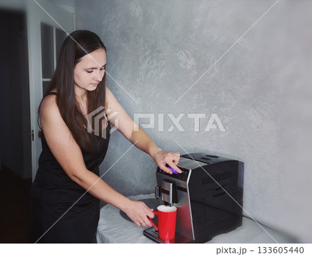 Woman Making Coffee with Automatic Espresso Machine. Young woman in black dress pressing button on modern coffee machine, placing red cup under spout. Morning ritual and home barista concept. 133605440