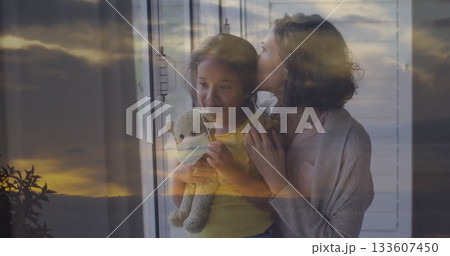 Mother embracing daughter wearing yellow top at home window, with gray teddy bear mask Mother embracing daughter wearing yellow top at home window, with gray teddy bear mask 133607450