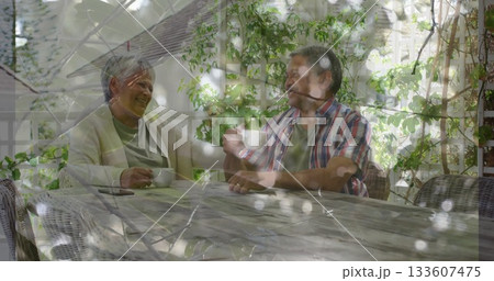 Sitting senior couple holding white coffee cups and checking phone on garden patio under pergola 133607475