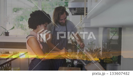 Cooking couple in aprons chopping vegetables and stirring pot at kitchen counter with sunset glow 133607482