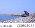 Man traveller is sitting on sea stone beach and browsing smartphone, side view. 133608029