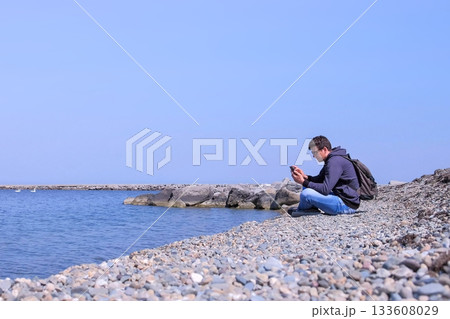 Man traveller is sitting on sea stone beach and browsing smartphone, side view. 133608029