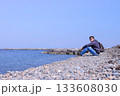Young man traveler sits on stone beach and looks at sea on spring vacation. 133608030