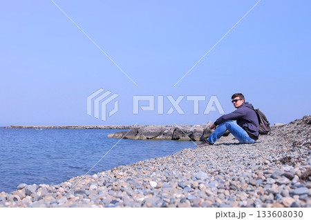 Young man traveler sits on stone beach and looks at sea on spring vacation. 133608030