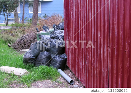 Black garbage bags near the fence of private house and an overturned trash can. Black garbage bags near the fence of private house and an overturned trash can. 133608032