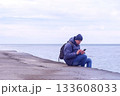 Man is typing a message on smartphone sitting at sea waterfront in overcast day. 133608033
