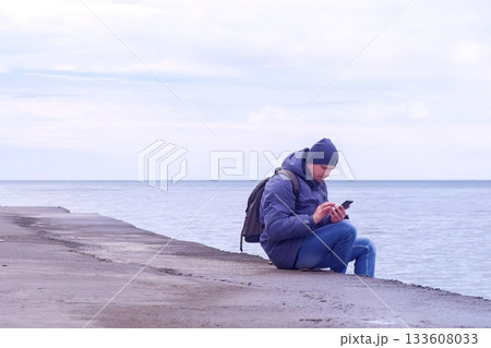 Man is typing a message on smartphone sitting at sea waterfront in overcast day. 133608033