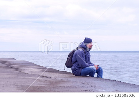 Man tourist sits on old sea waterfront and looks at sea in winter with phone. 133608036