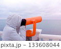 Woman in white jacket and hood looks in binoculars at sea waterfront with sea view. Side view. 133608044