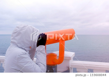 Woman in white jacket and hood looks in binoculars at sea waterfront with sea view. Side view. 133608044