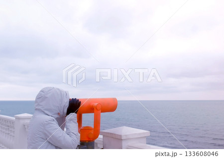 Woman in white jacket and hood looks in binoculars at sea waterfront with sea view. Side view. Woman in white jacket and hood looks in binoculars at sea waterfront with sea view. Side view. 133608046