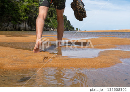 Hiker walking barefoot through water carrying shoes on Abel Tasman Track, NZ 133608592