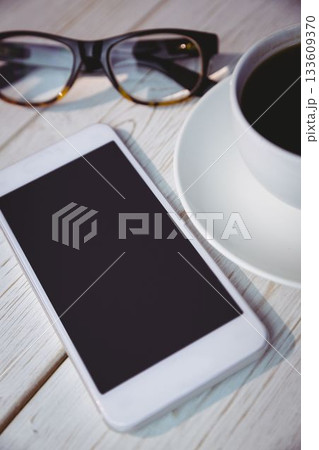 Showing pale wooden tabletop featuring white smartphone, black-framed eyeglasses and coffee cup 133609370