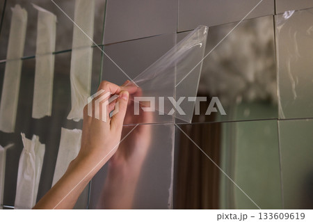 Removing protective layer to reveal clear glass, Carefully removing protective film from mirror to show pristine reflective surface in warm lit bathroom environment Removing protective layer to reveal clear glass, Carefully removing protective film from mirror to show pristine reflective surface in warm lit bathroom environment 133609619
