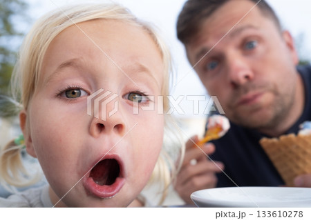 Dad and Little Girl Sitting in a Cafe, Eating Ice Cream and Looking Surprised at the Camera Dad and Little Girl Sitting in a Cafe, Eating Ice Cream and Looking Surprised at the Camera 133610278