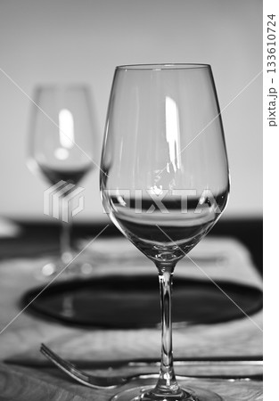 A refined black and white photograph of a table set for a formal meal, focusing on a clear wine glass in the foreground A refined black and white photograph of a table set for a formal meal, focusing on a clear wine glass in the foreground 133610724