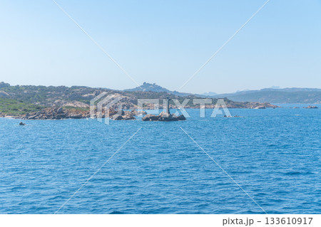 Calm blue water stretches toward a rocky shoreline with a solitary lighthouse standing on the stone edge. The landscape conveys a peaceful maritime atmosphere and a sense of open coastal space. Calm blue water stretches toward a rocky shoreline with a solitary lighthouse standing on the stone edge. The landscape conveys a peaceful maritime atmosphere and a sense of open coastal space. 133610917