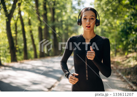 Focused front view, running, jogging. Young sportive skinny woman is in black fitness clothes outdoors 133611207