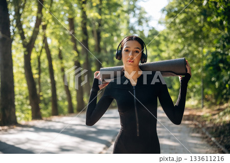 Skinny body type. Standing with yoga mat. Young sportive woman is in black fitness clothes outdoors Skinny body type. Standing with yoga mat. Young sportive woman is in black fitness clothes outdoors 133611216