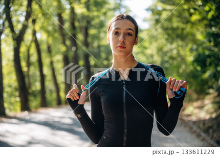 Under sunlight, with blue jumping rope, standing. Young sportive skinny woman is in black fitness clothes outdoors 133611229