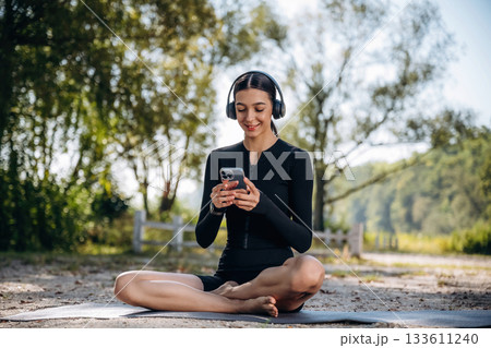 Relaxed, sitting on the yoga mat, break. Young sportive skinny woman is in black fitness clothes outdoors 133611240