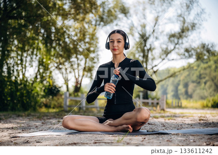 Relaxed, sitting on the yoga mat, break. Young sportive skinny woman is in black fitness clothes outdoors 133611242