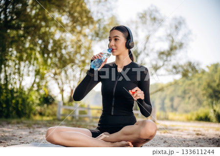 Rural place. Drinking water. Sitting. Young sportive skinny woman is in black fitness clothes outdoors 133611244