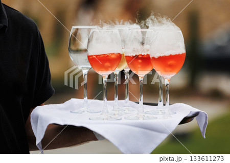 Close up of wine glasses with orange alcoholic cocktail decorates by bubbles with smoke steam by dry ice on tray with white cloth in hand of unrecognisable waiter. Wedding, birthday party outdoors Close up of wine glasses with orange alcoholic cocktail decorates by bubbles with smoke steam by dry ice on tray with white cloth in hand of unrecognisable waiter. Wedding, birthday party outdoors 133611273